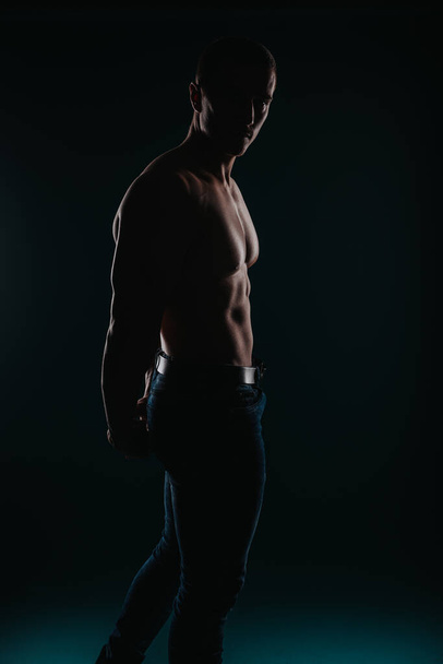 Homem bem construído e charmoso em pé na frente de um fundo escuro e flexionando seus músculos - Foto, Imagem