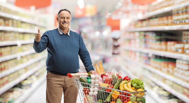 Mature man with a shopping cart at the supermarket gesturing thumbs up  - Valokuva, kuva