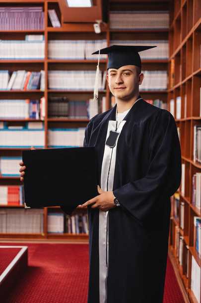 Genç ve mutlu mezun, kep ve cübbe giyiyor ve lisans diplomasını kütüphanede tutuyor. - Fotoğraf, Görsel