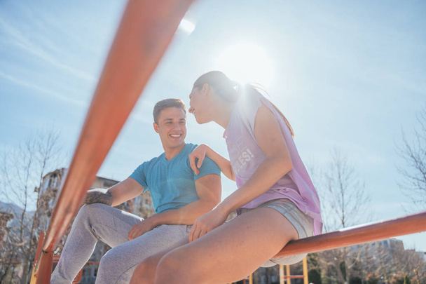 Sportliches Paar unterhält sich lustig und lächelt Sie machen eine Trainingspause, während sie auf einer waagerechten Metallstange sitzen. - Foto, Bild