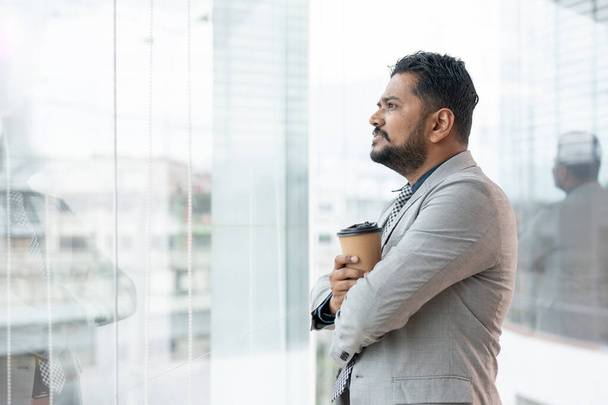 Úspěšný a odhodlaný indicko-asijský podnikatel nebo výkonný ředitel ve formálním obchodním obleku se dívá ven z budovy se šťastnou tváří a šálkem kávy v ruce. zadní pohled - Fotografie, Obrázek