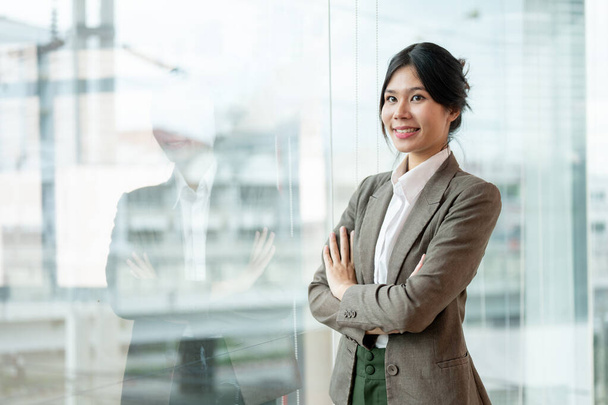 Ένα πορτραίτο μιας όμορφης Ασιάτισσας επιχειρηματία με επιχειρηματικό κοστούμι στέκεται με τα χέρια σταυρωμένα σε ένα διάδρομο γραφείου.. - Φωτογραφία, εικόνα