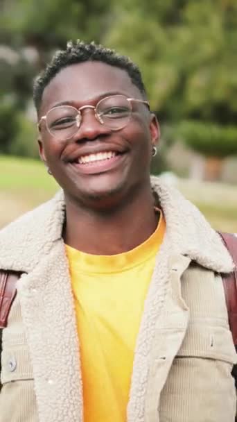Ritratto verticale ravvicinato di un adolescente afroamericano che guarda la telecamera sorridente e ride del campus universitario. Giovane studente intelligente in piedi all'aperto al college con zaino e googles. Alto - Filmati, video