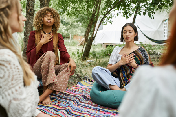 multiethnic women in stylish boho outfits meditating on meadow in retreat center - 写真・画像