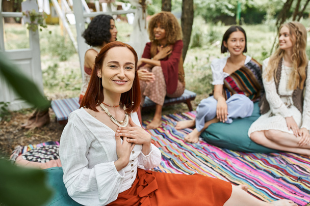 smiling redhead woman looking at camera near blurred multiethnic friends outdoors in retreat center - Foto, Imagem