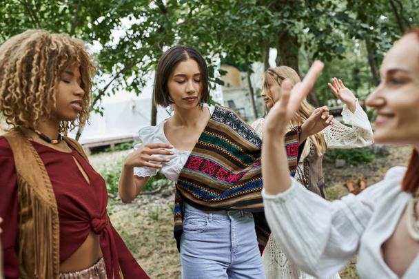 young woman in boho outfit dancing near interracial friends outdoors in retreat center - Zdjęcie, obraz