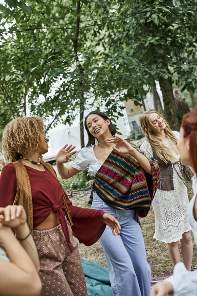 alegres mujeres multiétnicas en trajes boho bailando juntas al aire libre en el centro de retiros - Foto, Imagen