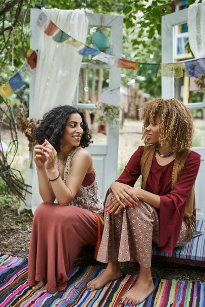 smiling multiethnic women in boho outfits talking while spending time outdoors in retreat center - Фото, зображення