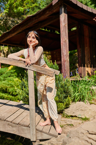 ευτυχισμένη και ονειρική Ινδή γυναίκα με παραδοσιακή ενδυμασία κοιτάζει μακριά σε ξύλινη γέφυρα στο πάρκο το καλοκαίρι - Φωτογραφία, εικόνα