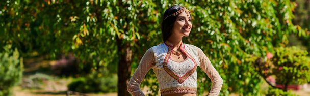 улыбающаяся индийская женщина в этнической одежде, стоящая с руками на бедрах и глядя в парке, баннер - Фото, изображение