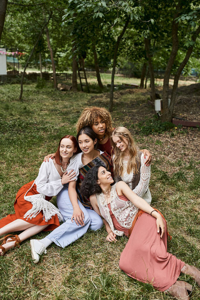 smiling interracial women in stylish boho outfits while sitting together outdoors in retreat center - Foto, Imagem