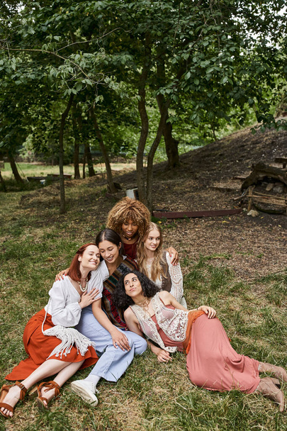 joyful interracial women hugging while sitting on meadow together in retreat center - Fotografie, Obrázek