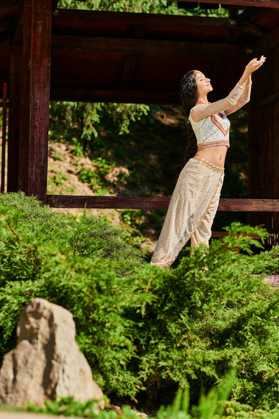 moda tradicional, mujer india complacida en ropa étnica de pie cerca de plantas verdes en el parque de verano - Foto, Imagen