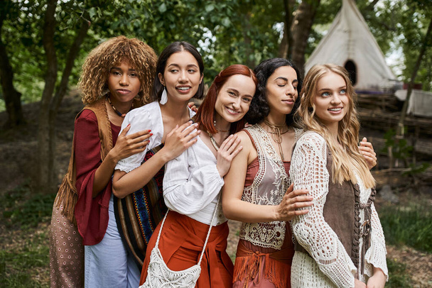 trendy and carefree interracial girlfriends in boho outfits hugging outdoors in retreat center - Photo, Image
