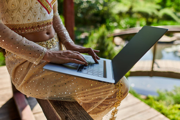 indiańska kobieta w eleganckim tradycyjnym stroju przy użyciu laptopa z pustym ekranem w letni dzień, widok częściowy - Zdjęcie, obraz
