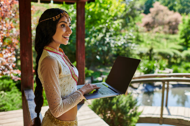 летний парк, улыбающаяся индианка в стильной этнической одежде держит ноутбук с чистым экраном - Фото, изображение