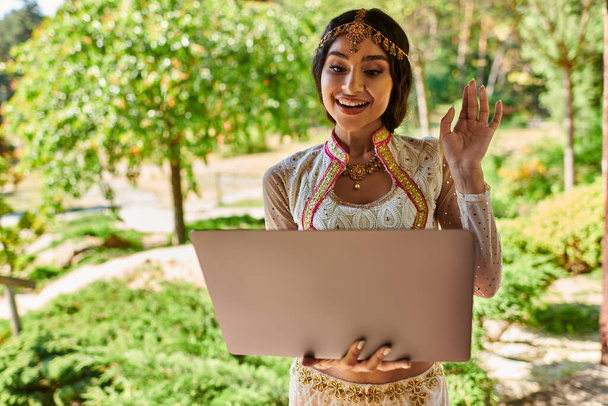 joyful indian woman in traditional attire waving hand during video call on laptop in summer park - Photo, Image
