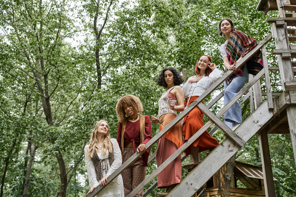 trendy and carefree multiracial women in boho style outfit on stairs of retreat center - Photo, Image