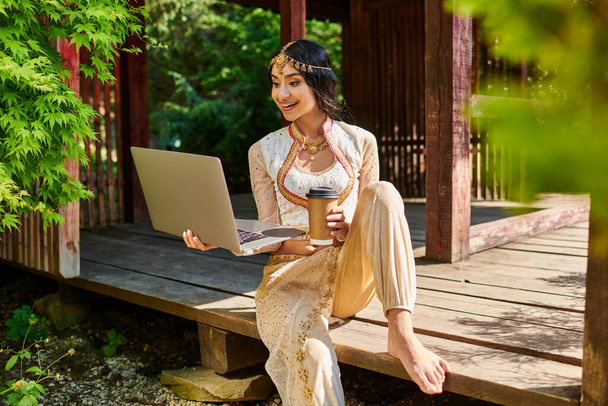 щаслива індійська жінка в етнічному одязі з ноутбуком і виймання кави в дерев'яному алькові в парку - Фото, зображення