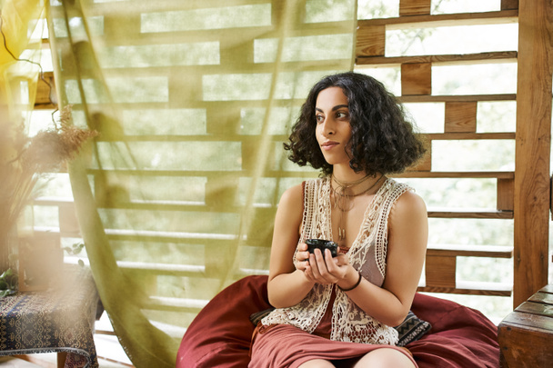 dreamy multiracial woman with tea cup sitting in bean bag in cottage, women retreat concept - Photo, Image
