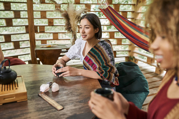 happy young woman in boho style clothes drinking tea at wooden table in retreat center - Photo, Image