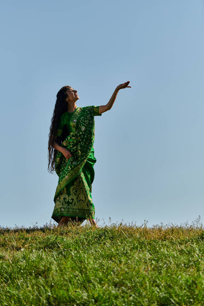 zomer, zonnige dag, vrolijke indiaanse vrouw in sari staand met uitgestrekte hand onder de blauwe hemel - Foto, afbeelding