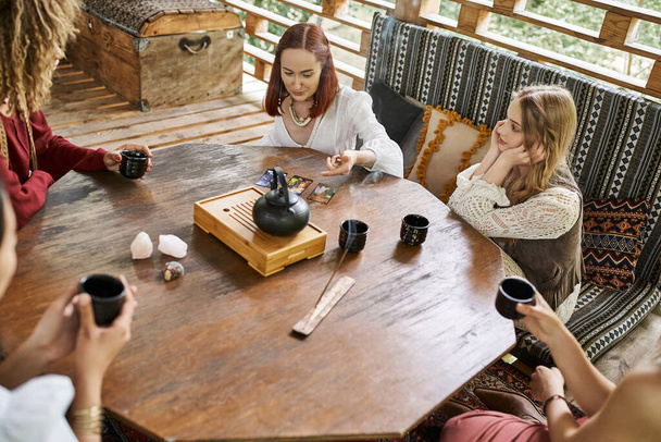 mujer leyendo cartas del tarot cerca de novias multiétnicas y té en mesa de madera en casa acogedora - Foto, Imagen