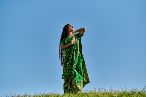 summer enjoyment, green field, indian woman in ethnic wear smiling with closed eyes under blue sky - Фото, изображение