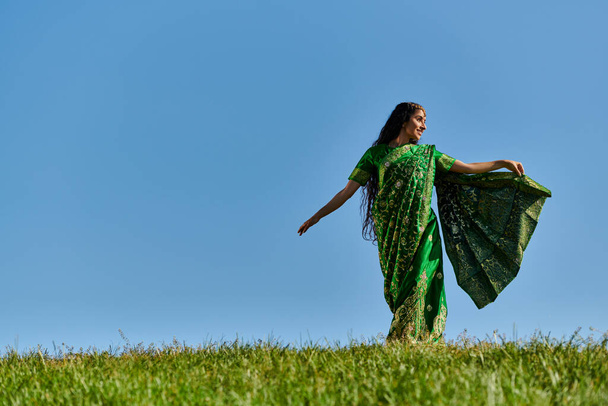 καλοκαιρινή μέρα, ανέμελη ινδική γυναίκα σε αυθεντικό φορούν περπάτημα στο πράσινο πεδίο κάτω από το γαλάζιο του ουρανού - Φωτογραφία, εικόνα