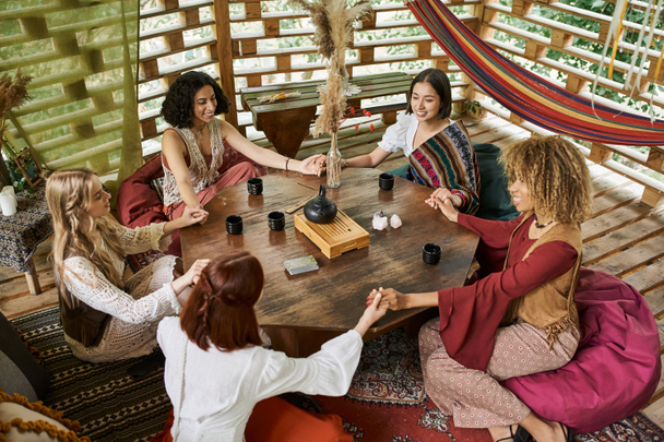 χαμογελαστές πολυφυλετικές φίλες που κρατιούνται χέρι-χέρι και διαλογίζονται σε ξύλινο τραπέζι με φλιτζάνια τσαγιού - Φωτογραφία, εικόνα