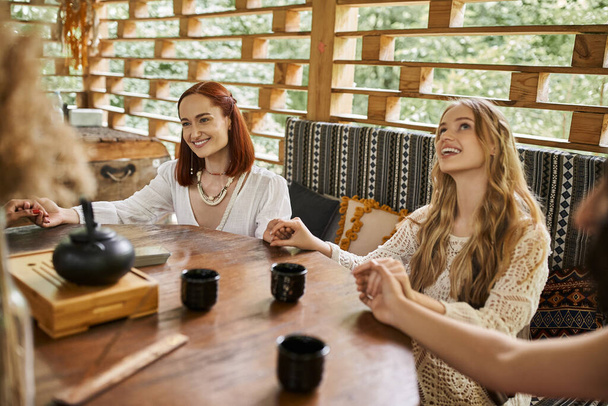 vrouwen retraite, zorgeloze vriendinnen mediteren en hand in hand nabij theekopjes op houten tafel - Foto, afbeelding