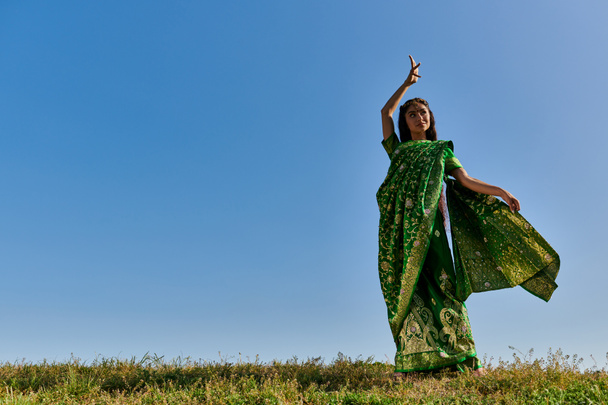 zomer dans van glimlachende indiaanse vrouw in authentieke sari in groen veld onder blauwe hemel - Foto, afbeelding