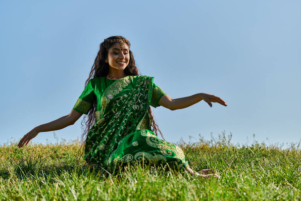 sonriente joven india mujer en verde sari sentado en la colina cubierta de hierba con el cielo azul en el fondo - Foto, imagen