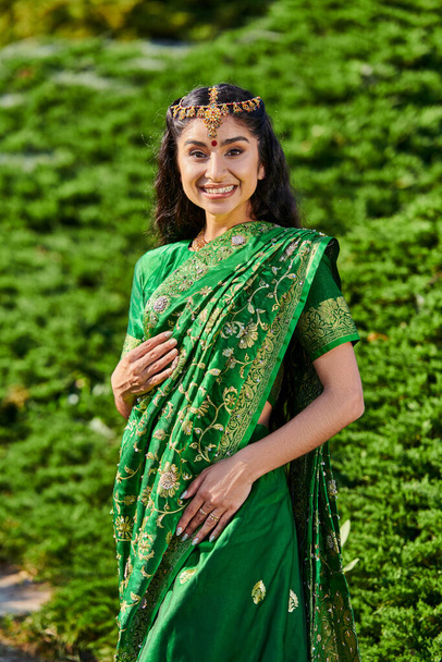retrato de joven mujer india alegre en sari tradicional y matha patti mirando la cámara en el parque - Foto, imagen