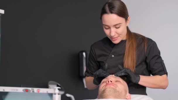 Homem europeu deitado em uma cama de spa recebendo massagem facial com óleo essencial aromático cuidados com a pele. Uma mulher massagista faz uma massagem de saúde em um salão de beleza - Filmagem, Vídeo