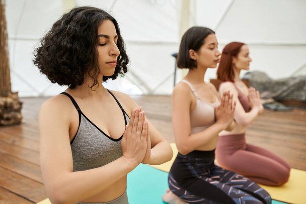 multiracial woman with closed eyes and praying hands practicing yoga with girlfriends, harmony - Fotografie, Obrázek