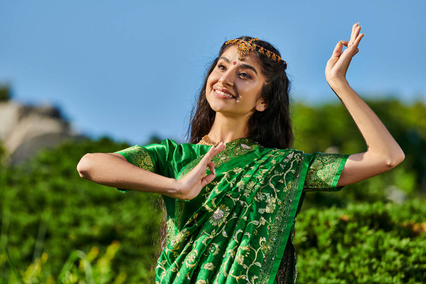 bonita jovem mulher indiana em sari tradicional dançando no parque borrado com céu azul no fundo - Foto, Imagem