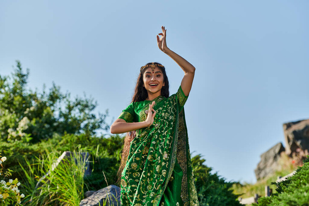 excited young indian woman in stylish green sari posing in summer park on background - Foto, afbeelding