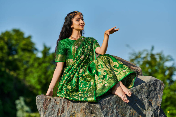 усміхнена і стильна босоніжка індійська жінка в сарі сидить на камені з блакитним небом на фоні - Фото, зображення