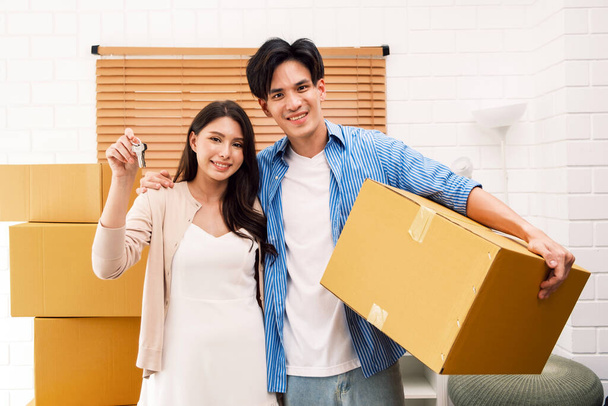 Nuori onnellinen aasialainen pari, jolla on kameran avaimet, kun mies pitelee pakettilaatikkoa uudessa kodissaan. Muuttoa, muuttoa omaan asuntoon, vuokrausta, kiinteistöjä. Uusi asunnon käsite. - Valokuva, kuva