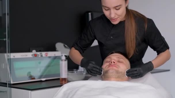 Молодой человек в спа, проходящий процедуру по уходу за лицом. Глиняная маска на лице человека. Косметолог делает массаж лица с увлажняющей маской, нанесенной на лицо - Кадры, видео