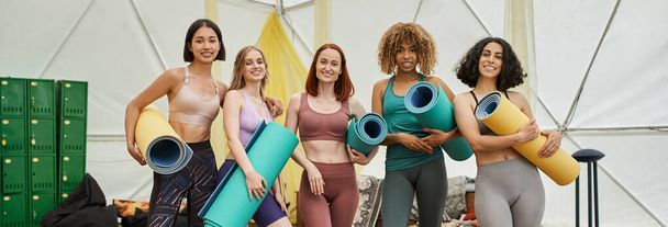 namoradas multiétnicas alegres em sportswear, com tapetes de ioga, olhando para a câmera, banner - Foto, Imagem