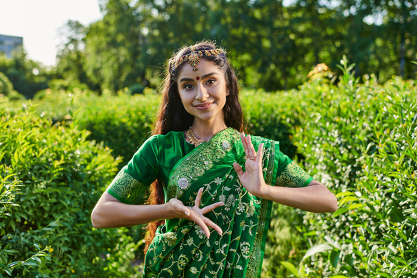 стильная молодая индианка в сари и бинди позирует и смотрит в камеру в парке - Фото, изображение