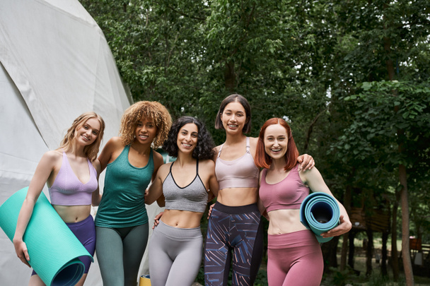 positive multiethnic girlfriends in sportswear holding yoga mats and smiling at camera, park - Photo, Image