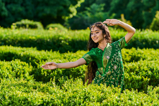 positive young indian woman in green sari and bindi posing near green plants in summer park - Fotó, kép