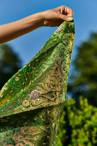 cropped view of young woman holding modern green sari with pattern near blurred plants outdoors - Photo, Image