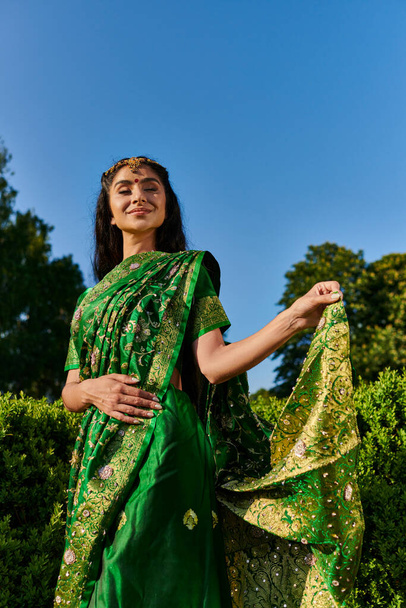 cheerful and modern young indian woman touching sari with pattern near green plants in park outdoors - Photo, Image