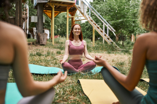 joyful woman with closed eyes meditating in park of retreat center near blurred girlfriends - Foto, afbeelding
