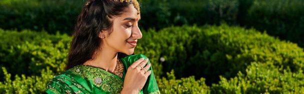 side view of elegant young indian woman in green sari standing near blurred plants in park, banner - Photo, Image