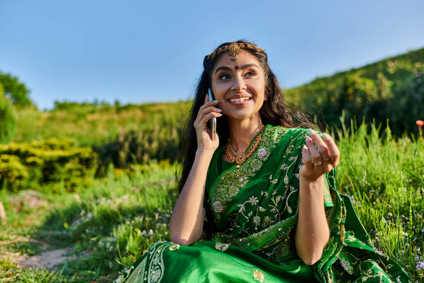 Moda elbisesi içinde neşeli, genç Hintli bir kadın yazın çimenlerde otururken akıllı telefonuyla konuşuyor. - Fotoğraf, Görsel